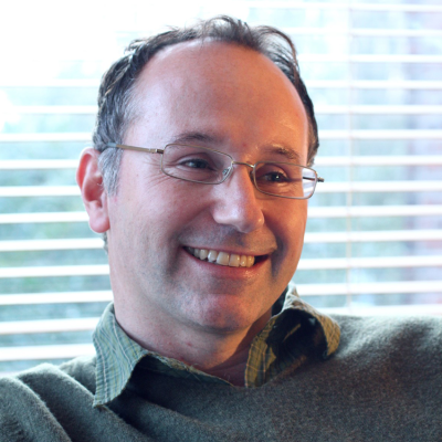 A white man with classes and a green jumper is sitting in front of a window smiling.